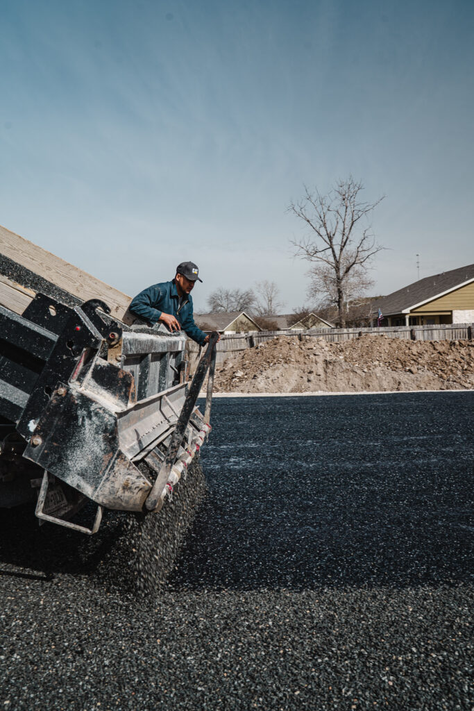 A worker operates an asphalt paving truck to lay fresh asphalt on a residential driveway, with gravel stockpile and houses visible in the background.
