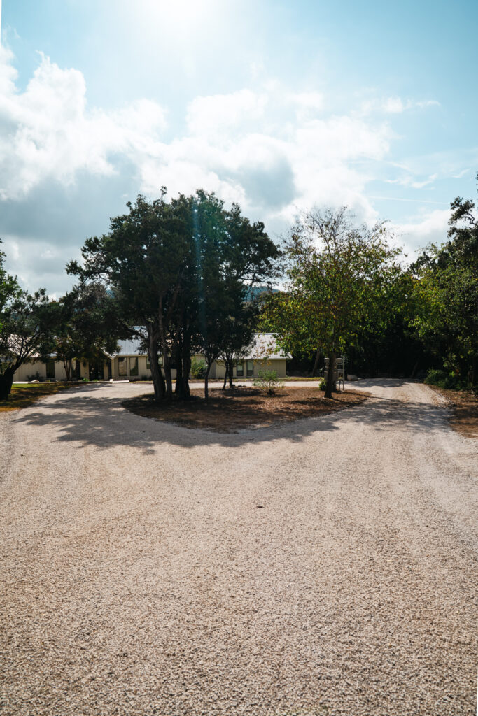 Faded chip seal surface on private driveway in Mountain Home TX: a rustic gravel driveway with mature trees and ranch-style