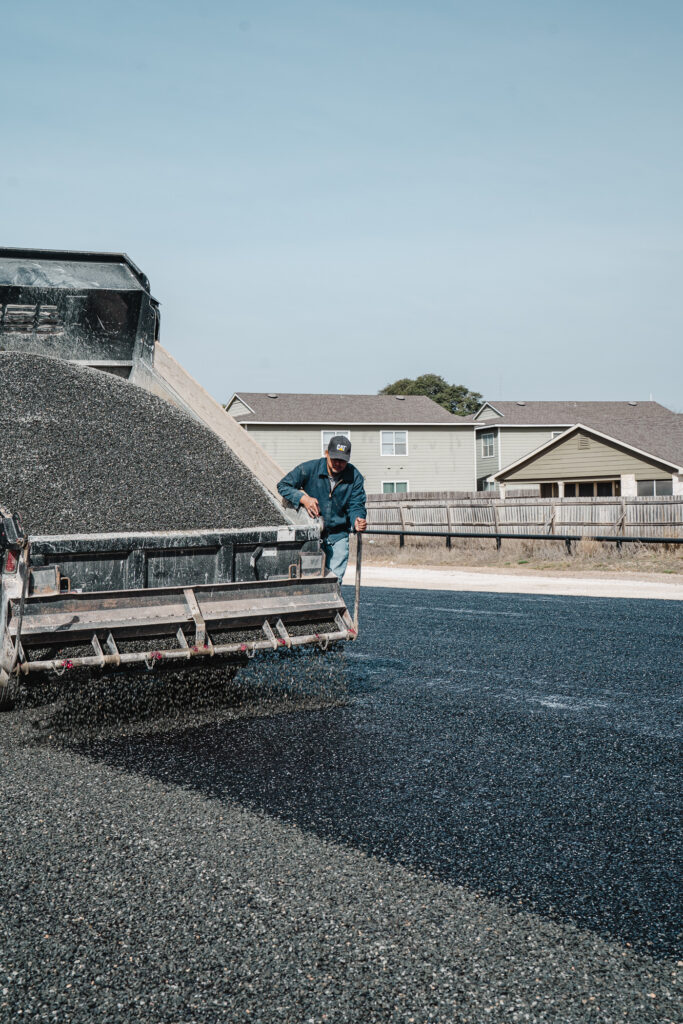 Fog seal application preventing oxidation on driveway in Lake Medina Shores TX: a worker operates an asphalt paving machine