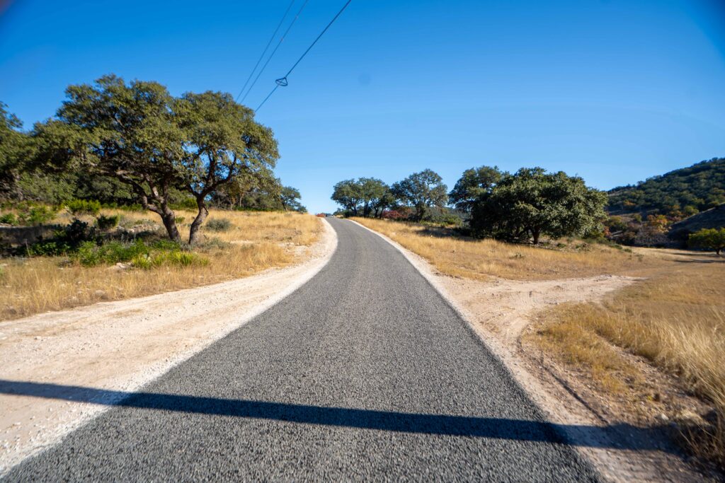 A newly paved asphalt road stretching through a rural landscape with scattered oak trees and golden dry grass on both sides under clear blue sky.