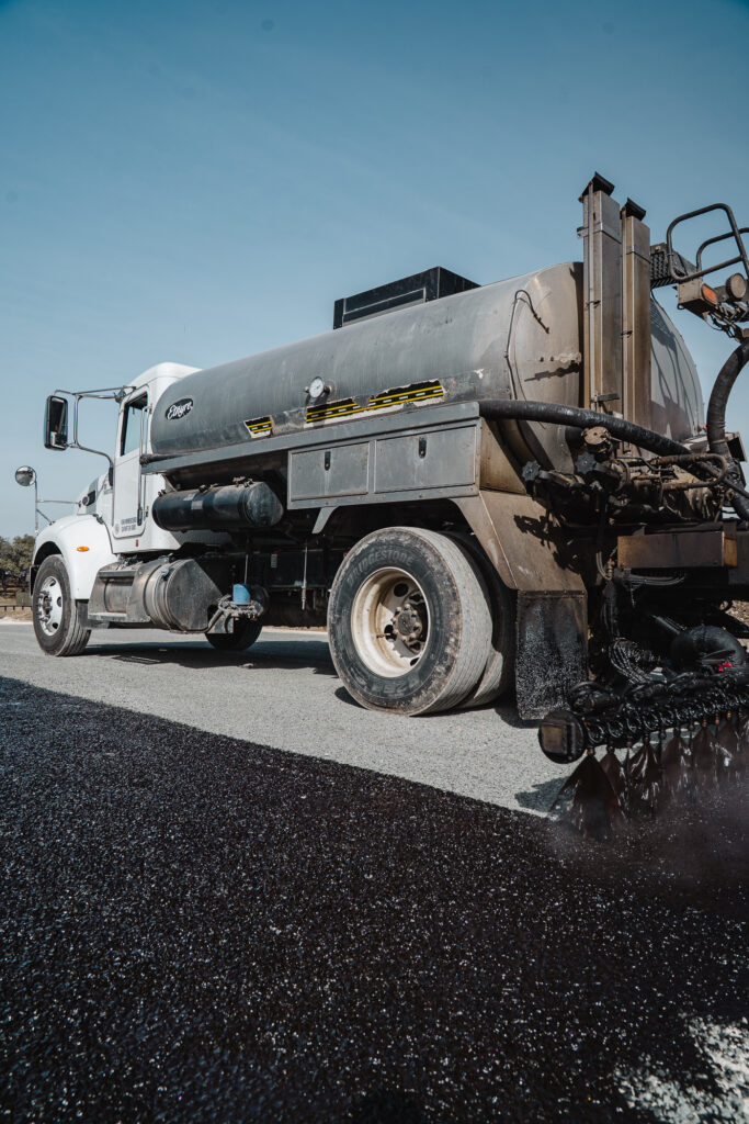 White streaking and calcium deposits on chip seal surface in Grape Creek TX: a white Isuzu vacuum truck with large storage